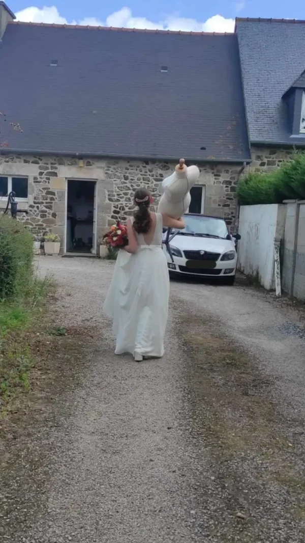 Marie en robe de mariée marchant de dos avec son mannequin sur l'épaule
